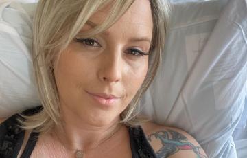 young woman with blond hair and long eyelashes lying in hospital bed wearing a black sleeveless top showing a port and a tattoo