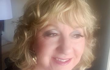 hazy photo of middle-aged woman with blond hair gray eyeshadown pink lipstick and gold earrings and necklace in a black dress
