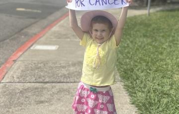 little white girl in a yellow shirt and pink skit with butterflies on it wearing a hat and holding up a sign that says honk if you want a cure for cancer