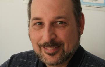 middle aged white man with a beard and mustache wearing a navy blue shirt sitting in front of a world map