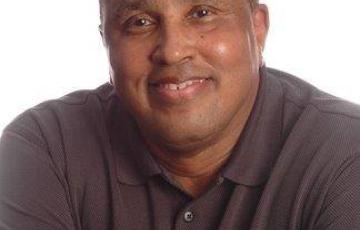 older black man smiling wearing a burgundy colored shirt with his arms crossed