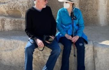 older white couple sitting in front of stones man wearing burgundy ball cap and black shirt and woman wearing blue shirt face maske and straw hat