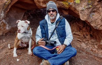 older white man with beard and mustache wearing a knit cap and puffy blue vest holding a camera sitting by his dog