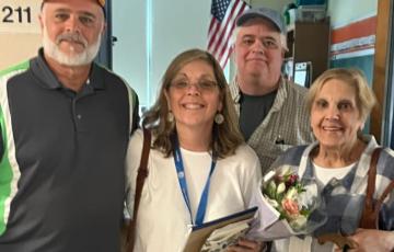two older men wearing ball caps with a woman wearing glasses and dangly earrings holding a bouquet of flowers and their mom on the right and an American flag in the background
