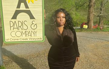 young black woman wearing a black dress with gold hoop earrings and necklace standing by a sign that says Paris and Company