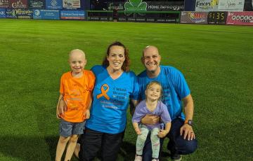 little white boy who is bald wearing an orange t-shirt and blue shorts next to his mom sister and dad wearing blue and orange tshirts on a ball field