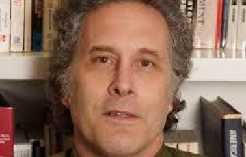 middle aged white man with graying sideburns and a green shirt and red tie sitting in front of a bookcase