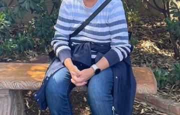 middle aged white woman short brown hair sitting on a bench and wearing a blue striped shirt and jeans with a jacket tied around her waist