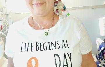 middle aged white woman wearing a colorful headscarf and white t-shirt with an orange ribbon on it sitting on a hospital bed