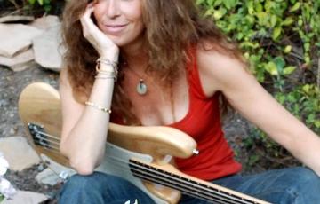 middle aged white woman with long brown hair wearing and orange tank and jeans with a guitar on her lap sitting in a garden