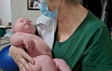 middle aged white woman with short hair and glasses wearing a face mask and a green shirt holding a baby