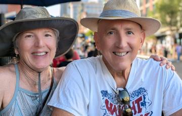 older white couple both wearing hats and smiling man wears white t-shirt with sunglasses hanging from the neck