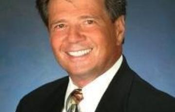 older white man headshot smiling wearing a brown suite white shirt and brown and blue tie