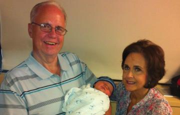 older white man with glasses and blue striped shirt holding a baby with an older woman with dark hair and a floral blouse