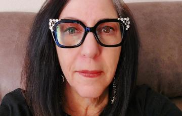 older white woman with dark long hair wearing black chunky glasses with bling on them and a black top