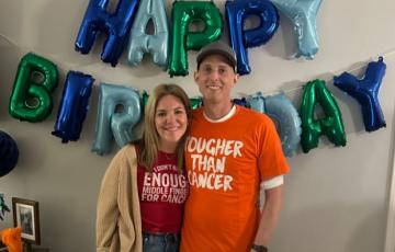 young white couple standing in front of a birthday balloon sign she has blond hair and burgundy t-shirt on he is wearing a ball cap and orange t-shirt