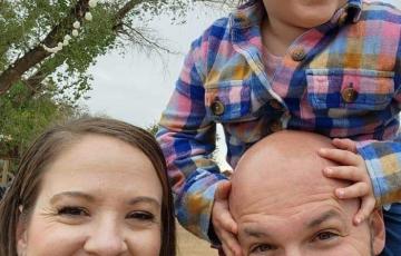 young white couple with their daughter outside little girl has on plaid shirt dad is bald with a mustache and beard mom has long brown hair