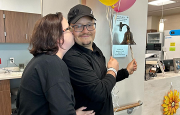Hispanic couple both dressed in black se is kissing him as he rings the bell and holds balloons