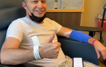 middle age white man with short hair, a black mask pulled down, a blue sleeve on his arm giving a thumbs up wearing white shirt with plaid shorts