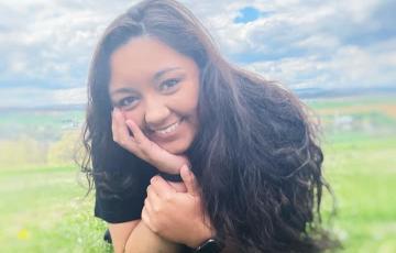 smiling young hispanic girl with long dark hair lying in the grass head propped on hand wearing black shirt and watch
