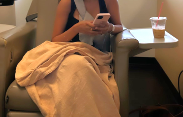 young ethnic girl sitting in a hospital chair wearing a yellow ball cap and a black sleevelss tshirt holding a pink phone with a blanket on her lap