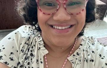 young hispanic woman with dark curly hair and red and white glasses wearing a white shirt with black flowers