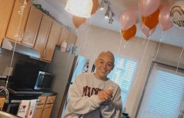 young white woman bald with a nose ring wearing a sweatshirt and standing in a kitchen with balloons and a bundt cake