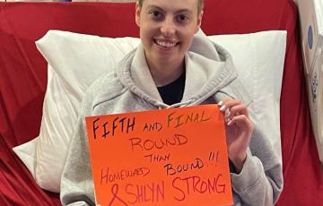 young white woman in a hospital bed with short hair wearing a gray hoodie and holding an orange sign about fifth round of chemo