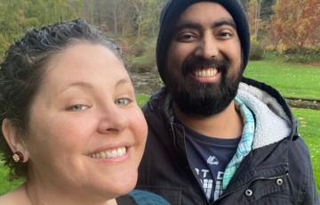 young white woman in blue sweater holding coffee with young black man with beard and mustache wearing a knit cap and blue jacket holding a cup