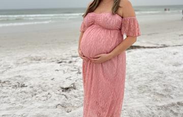 Holly young white woman with long hair and very pregnant wearing a pink off the should lace dress standing on a beach