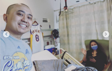 Nino Asian man in hospital room wearing blue tshirt standing by a Kobe Bryant jersey with his wife sitting in a chair waving and wearing a blue mask and dark shirt