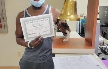 middle aged black man wearing LA ballcap and gray and white tank top and gray sweat pants holding a certificate and ringing a bell.