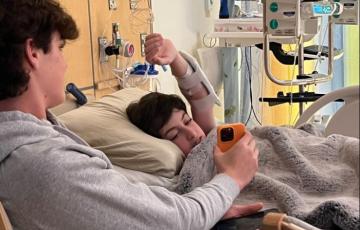 young white teen boy with dark hair lying in a hospital bed with his arm extended over his head looking at an orange phone being held by another teen boy