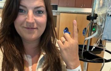 young white woman with long brown hair wearing a black shirt and white jacket sitting in a hospital room holding up her pointer finger