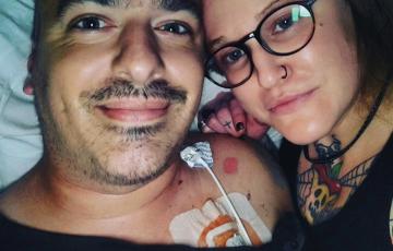 younger white man with bushy eyebrows and mustache wearing a black shirt with a port lying next young white woman with black glasses and a black nose ring with a tattoo on her finger and neck