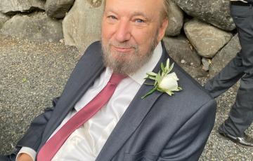 Gary older white man with blue eyes and thinning hair and gray beard and mustache leaning on rocks wearing a blue suite, mauve tie and a rose boutonaire