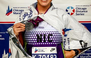 Nicolas smiling middle aged hispanic woman with dark hair pulled up in a bun wearing orangish sunglasses and a Team In Training Shirt holding a medallion in front of a Chicago Marathon banner.jpg