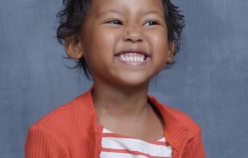 Sofia school picture of pre-K black girl with short curly hair and a big smile wearing and orange and white striped top under an orange sweater with a zipper