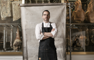 Steve young white man with short hair glasses and a beard and mustache wearing a white chef's coat under a denim apron standing in front of a canvas that is front of windows showing meats being cured