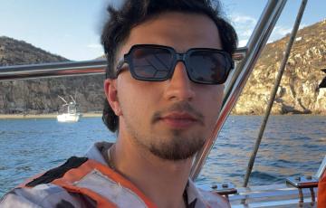 Steven young white male with dark hair and light mustache and beard wearing sunglasses and life vest over white and black shirt standing on a boat