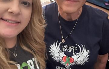 Susanne middle aged white woman with a bald head wearing hoop earrings and a heart necklace and a SCT Survivor t-shirt standing next to a young woman with a nose ring and Support Squad t-shirt