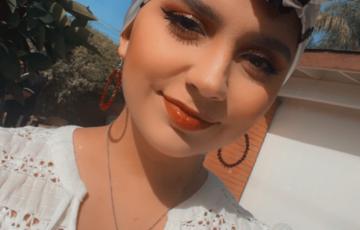 Yesenia young hispanic woman with blue and black headscarf on hoop earrings and a heart necklace wearing a white lacey blouse