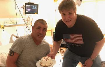 Greg two middle aged white men, one sitting on a hospital bed wearing a grey t-shirt and black shorts the other wearing a flag shirt and jeans both holding a cake