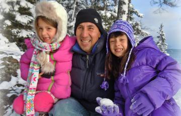 Joaquin middle aged hispanic man wearing navy beanie and ski jacket with two hispanic girls one in a pink jacket and one in a purple jacket holding a snowball in front of snow covered trees