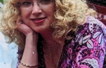 middle aged white woman with curly blonde hair and glasses with her chin resting on her hand wearing a pink and purple print shirt