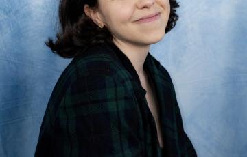 Crystal young hispanic woman with dark hair and gold hoop earrings smiling and wearing a blue and plaid top with blue photo background