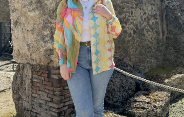 Eva young white woman with reddish hair sunglasses on her head in a white tshirt under a multi color shirt with blue jeans standing in front of a stone wall.