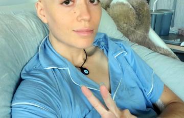 Josie young white woman bald with an orange tatoo and wearing blue pajamas in a hospital bed giving the peace sign