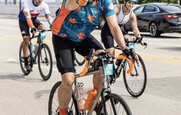 Michael older white man with white beard and mustache wearing an orange bike helmet and blue and orange shirt riding a bike while giving a thumbs up