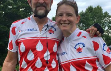 Paul older white gentleman with beard and mustache wearing a bike helmet and sunglasses with younger white female with freckles and sunglasses on her head both wearing Scenic Shore 150 shirts